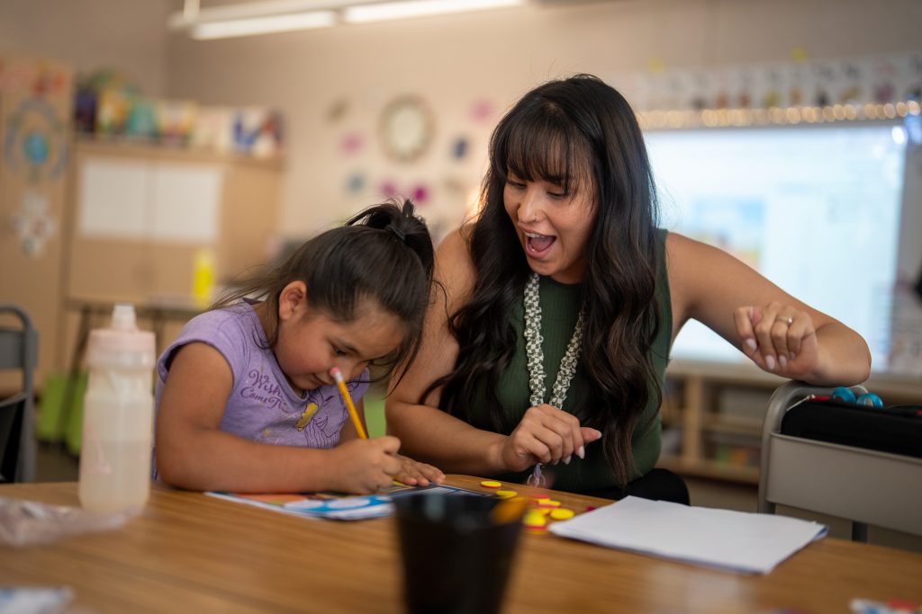Una profesora y una niña realizan una actividad creativa sentadas a una mesa; ambas sonríen y están concentradas en su trabajo.