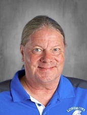 A middle-aged man with long hair wearing a blue polo shirt, smiling against a gray background.