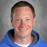 Smiling man with short brown hair wearing a blue hoodie over a gray background.