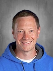 Smiling man with short brown hair wearing a blue hoodie over a gray background.