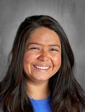 Smiling woman with long dark hair, wearing a blue shirt, set against a gray background.