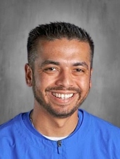 Smiling man with short hair, wearing a blue shirt, set against a gray background.