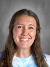 Smiling woman with long brown hair, wearing a tie-dye shirt, against a light gray background.