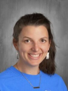 Smiling woman with dark hair in a blue shirt, wearing large earrings, against a gray background.