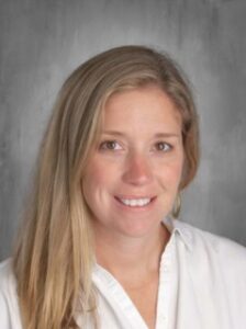 A smiling woman with long blonde hair is wearing a white shirt, posing against a gray background.
