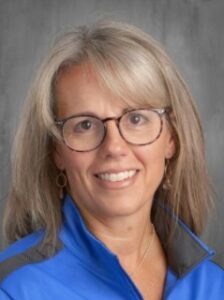 A smiling woman with glasses and shoulder-length gray hair, wearing a blue jacket, against a gray background.