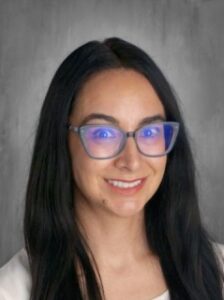Headshot of a woman with long black hair and glasses, smiling against a gray background.