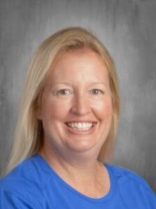 Smiling woman with long blonde hair wearing a blue shirt, set against a gray background.