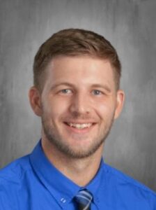 Smiling man with short light brown hair, wearing a blue shirt and tie, against a gray backdrop.