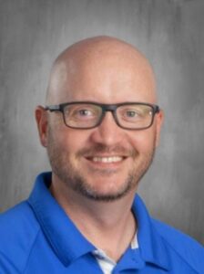 A smiling man with glasses and a bald head, wearing a blue collared shirt, against a gray background.