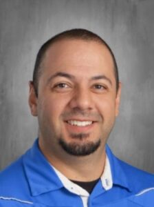 A smiling man with short dark hair and a goatee, wearing a blue shirt, against a gray background.