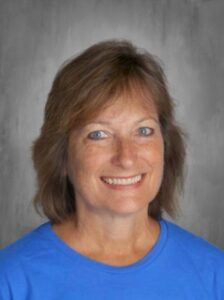 A smiling woman with shoulder-length brown hair, wearing a blue shirt, against a gray background.