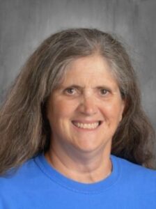 Smiling woman with long gray hair, wearing a blue shirt, against a blurred gray background.