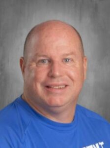 Smiling man with a receding hairline wearing a blue shirt, set against a gray background.