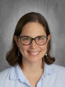 Smiling woman with shoulder-length dark hair, wearing glasses and a light blue shirt, against a neutral background.