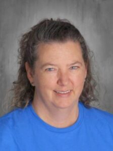 A smiling woman with curly hair wearing a blue shirt, set against a gray background.
