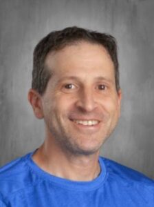 Headshot of a smiling man with short brown hair, wearing a blue shirt, against a gray background.