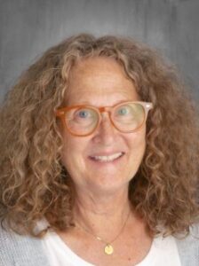 Woman with curly, brown hair and orange glasses, smiling against a gray background.