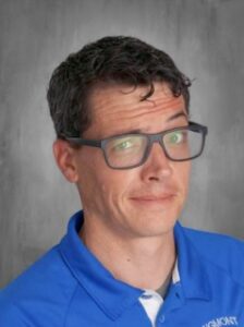 A man with short curly hair and glasses, wearing a blue shirt, smiles slightly, set against a gray background.