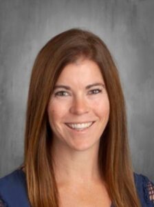 A woman with long brown hair smiles in front of a textured gray background, wearing a blue top.
