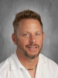 Portrait of a man with short, light brown hair and a beard, wearing a white shirt and a necklace, smiling at the camera.