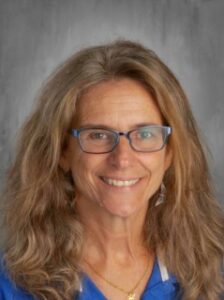 Smiling woman with long, wavy brown hair, wearing glasses and a blue shirt, set against a neutral background.