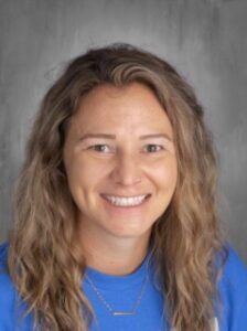 Smiling woman with wavy hair, wearing a blue shirt, against a gray background.