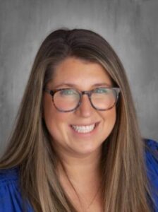 Smiling woman with long brown hair, wearing glasses and a blue shirt, against a grey background.