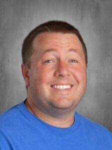 A smiling man with short brown hair, wearing a blue t-shirt, against a gray background.