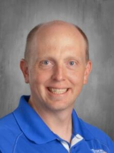 Portrait of a smiling man with short hair, wearing a blue polo shirt, set against a gray background.