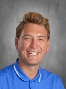 Smiling man with light brown hair wearing a blue polo shirt, set against a gray background.