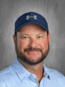 Smiling man with a beard wearing a blue cap and a light blue shirt, set against a neutral background.
