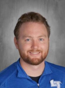 Smiling man with a beard, wearing a blue jacket, against a gray background.