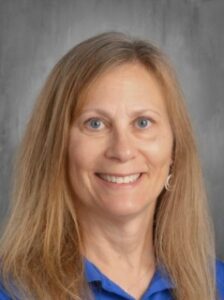 Smiling woman with long brown hair wearing a blue shirt, against a neutral gray background.