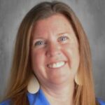 Smiling woman with long brown hair, wearing blue attire and large earrings, set against a neutral background.