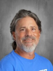 Smiling man with long gray hair and a beard, wearing a blue shirt, against a gray background.