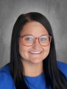 Smiling woman with long dark hair wearing glasses and a blue shirt, against a gray background.