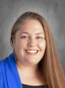 A smiling woman with long brown hair, wearing a blue jacket, against a neutral gray background.