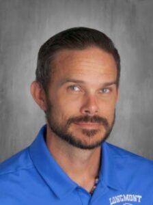 A man with short hair and a beard smiles, wearing a blue polo shirt with "LONGMONT" on it, against a gray background.