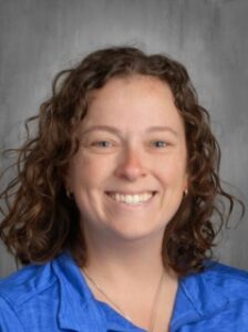 Smiling woman with curly hair wearing a blue shirt, against a gray background.