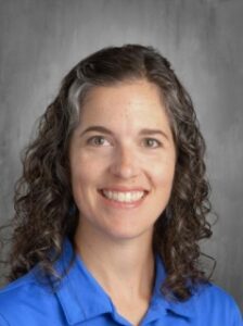 Portrait of a woman with curly hair wearing a blue shirt, smiling in front of a gray background.