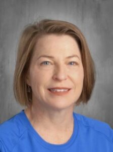A woman with short brown hair, wearing a blue shirt, smiles at the camera against a gray background.