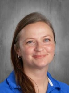 Smiling person with long brown hair, wearing a blue shirt, against a gray background.