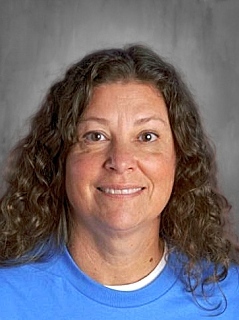 A woman with curly brown hair smiles, wearing a blue shirt, against a neutral background.