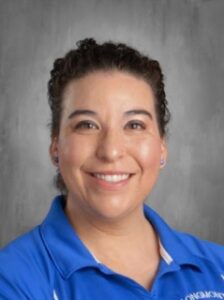 Smiling woman with curly dark hair, wearing a blue polo shirt, against a gray background.