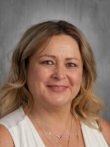 A close-up portrait of a woman with wavy, shoulder-length hair and a friendly smile, wearing a white top.