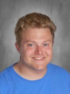Smiling young man with short, wavy hair, wearing a blue t-shirt, set against a gray background.