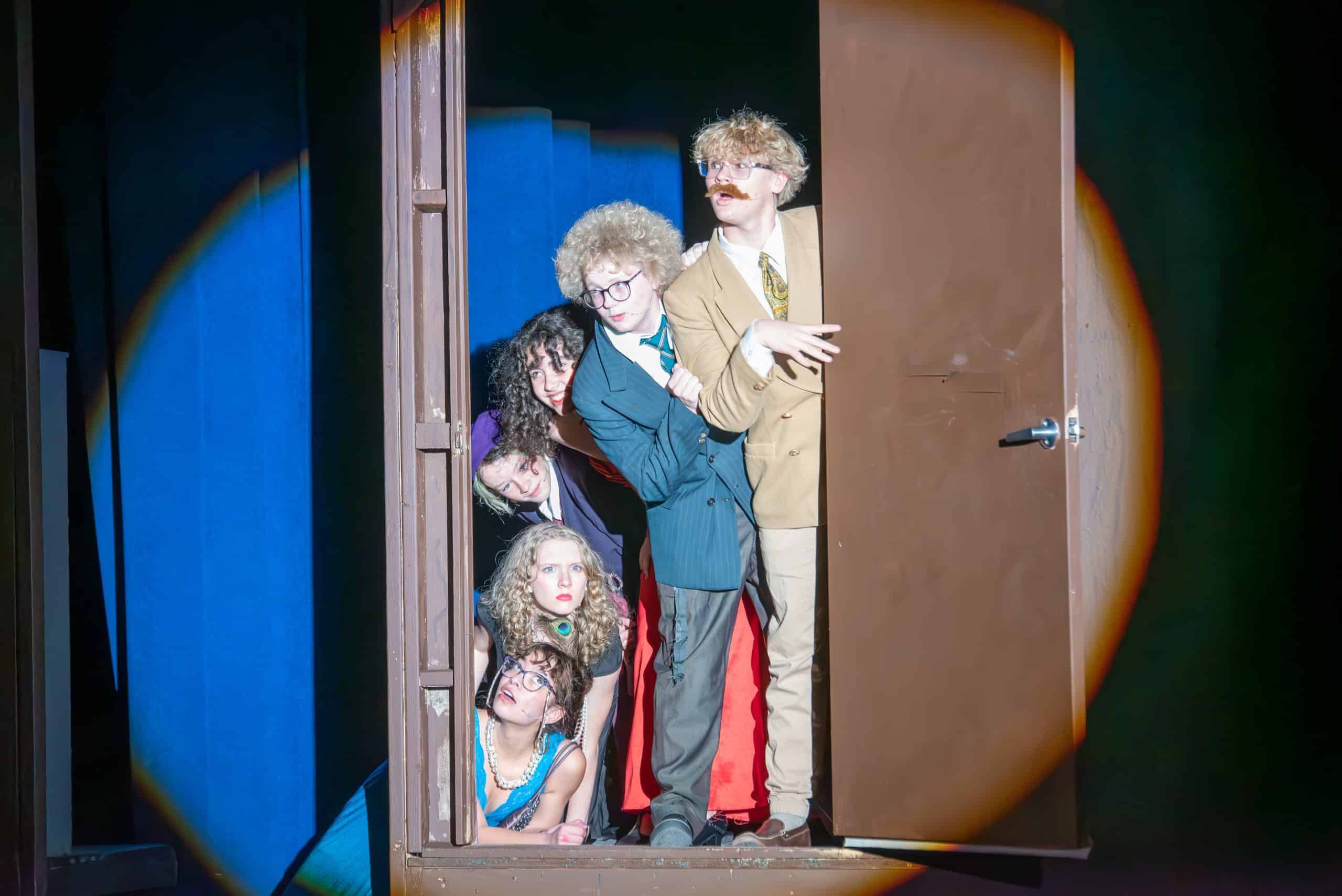 Seis estudiantes del club de teatro de Longmont se asoman por una puerta en el decorado del escenario. Una chica está tumbada en el suelo y los demás estudiantes están apilados encima de ella, de sentados a de pie.