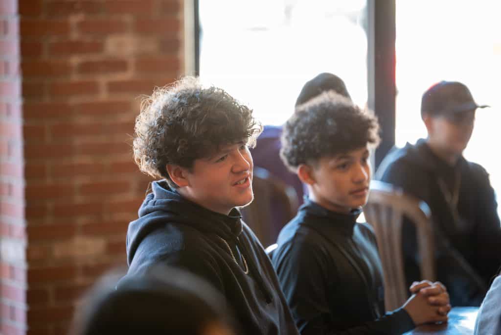 Two high school males with curly hair listening to a speaker at a local restaurant. Sitting in chairs with brick wall and windows behind them.