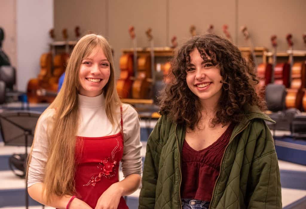 Two high school females, Madison Hatfield (left) and Brooklyn Rowinski (right) standing side-by-side smiling for camera. A row of cellos is in the background behind them. 